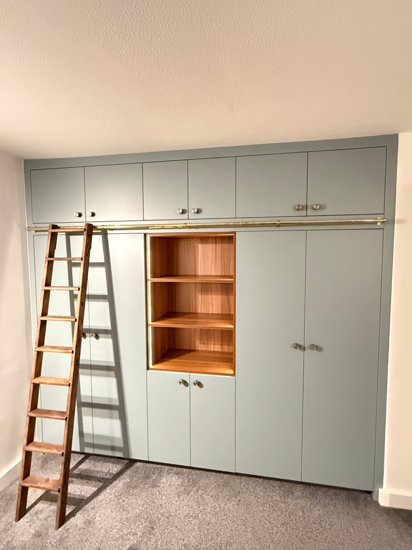Floor-to-ceiling sage green fitted wardrobes with oak open shelving and a library ladder
